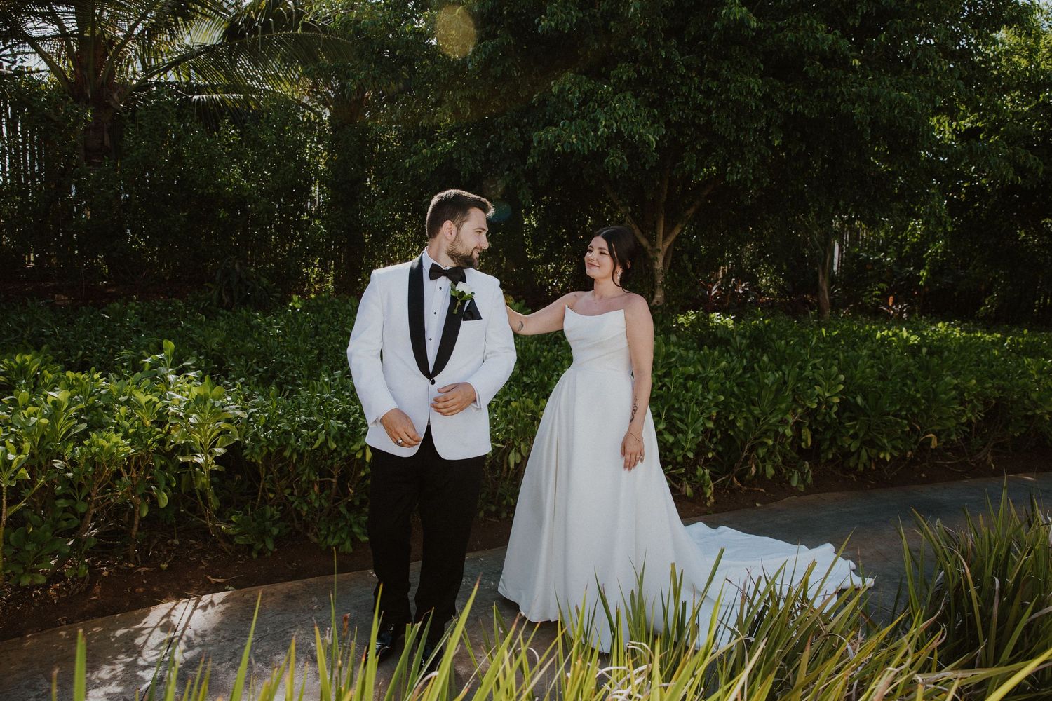 A bride in a flowing white gown and groom in white tuxedo jacket stand together on a garden path during sunset.