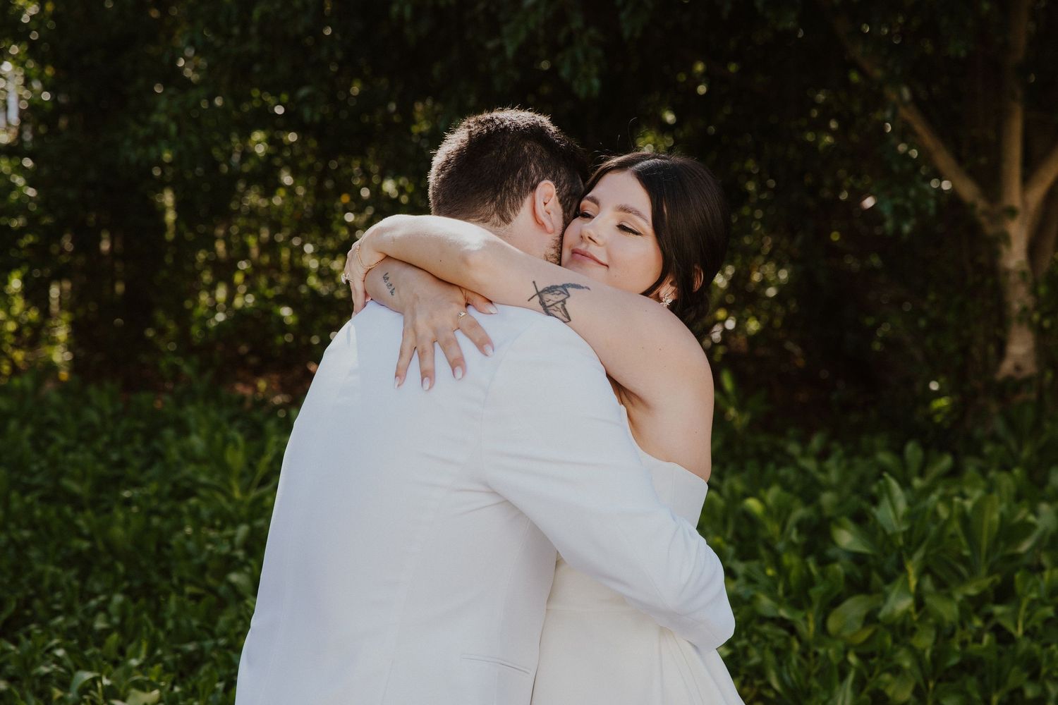 A romantic wedding couple embrace outdoors in a garden setting with lush green foliage in the background.