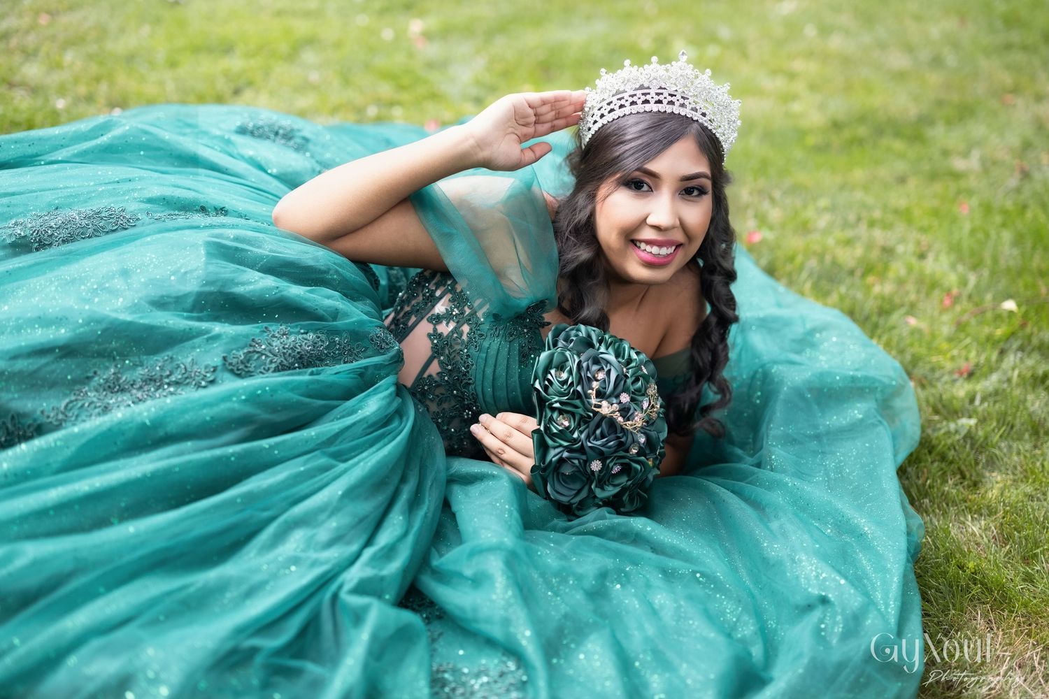 A person in a flowing teal quinceañera dress spins gracefully on grass while wearing a sparkling tiara.
