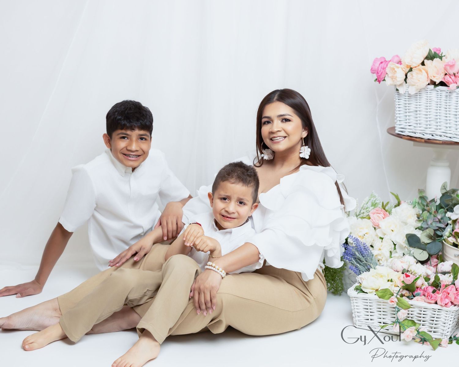 A family sits together in a bright studio setting with floral decorations and baskets in the background.