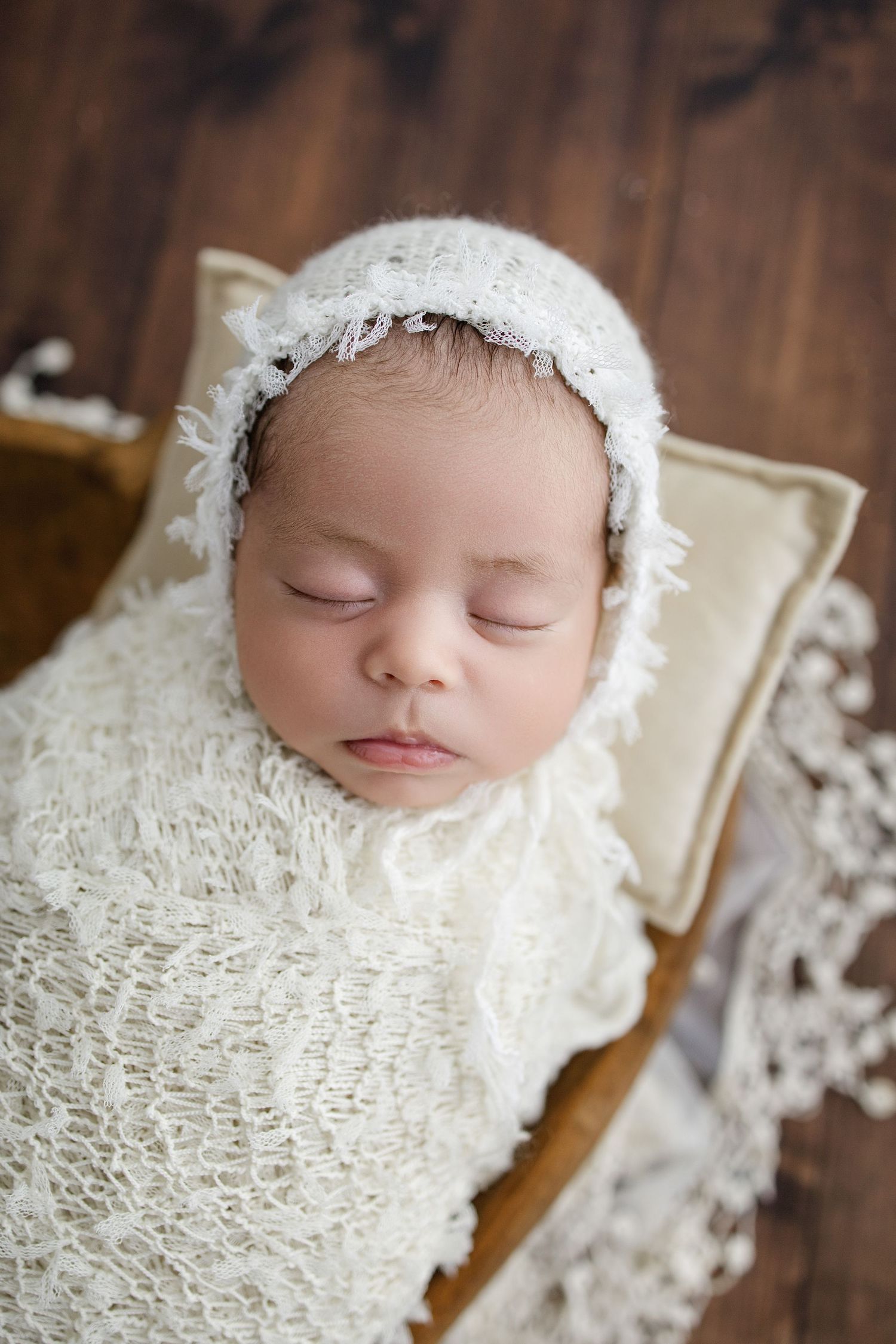 Peaceful sleeping newborn wrapped in white swaddle rests in cozy beige cushioned basket on wooden floor.