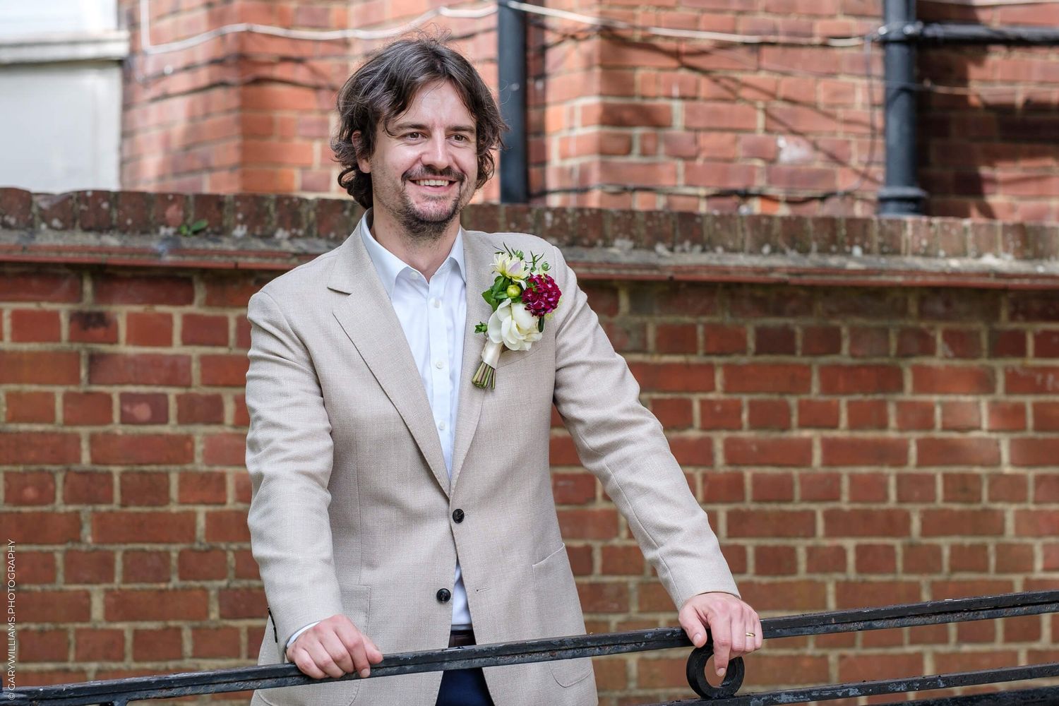 A groom in a beige suit stands along a brick wall on a wedding day.