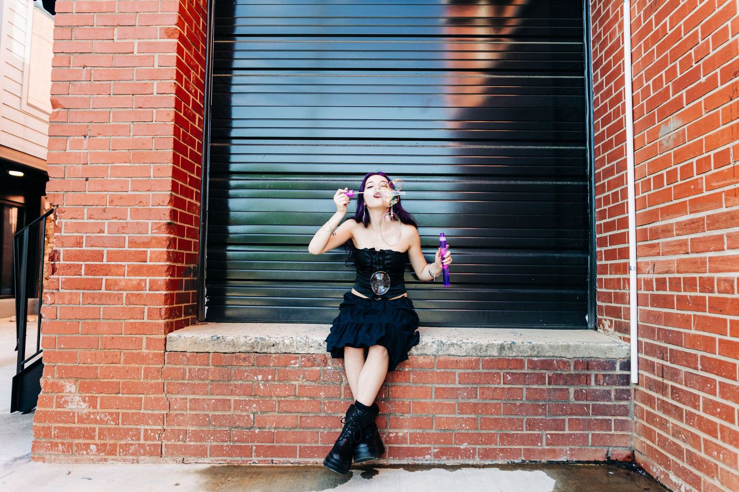 Person in black dress and boots poses against industrial green door and brick wall backdrop in urban setting.