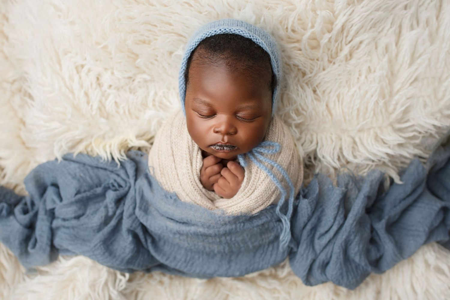 Peaceful sleeping newborn baby wrapped in cream and blue fabric laying on soft white fur blanket. south sudanese newborn photographer.