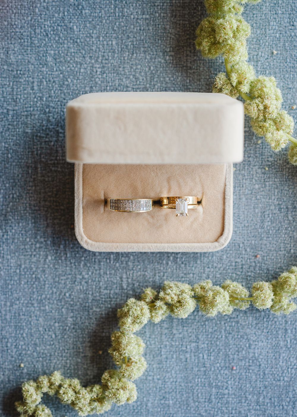 Wedding ring set displayed in a light beige velvet ring box on denim blue fabric with dried flowers.