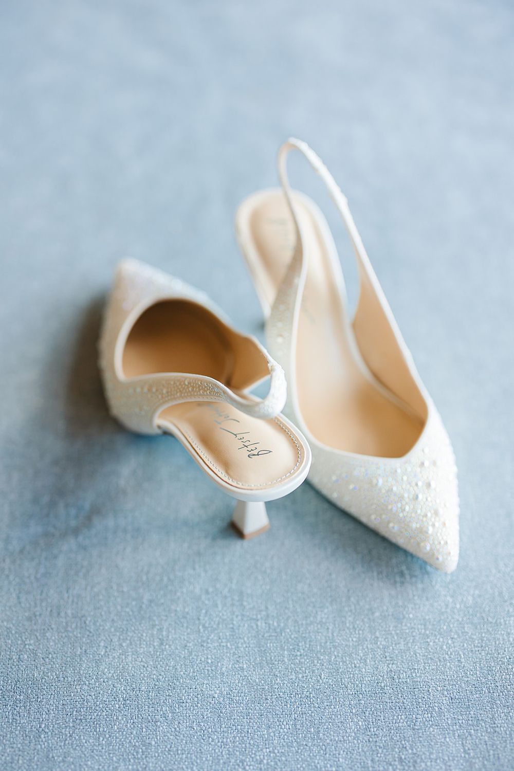 Elegant white lace bridal heels with delicate detailing photographed on a soft blue-gray background.