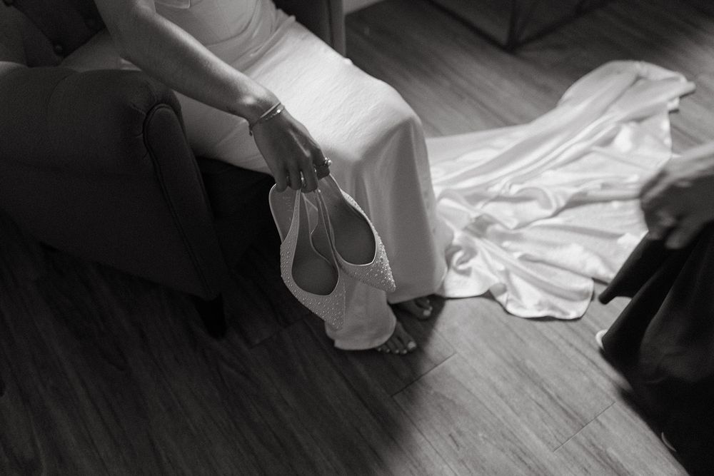 Black and white photo showing the hem and train of an elegant white wedding dress with matching heels.