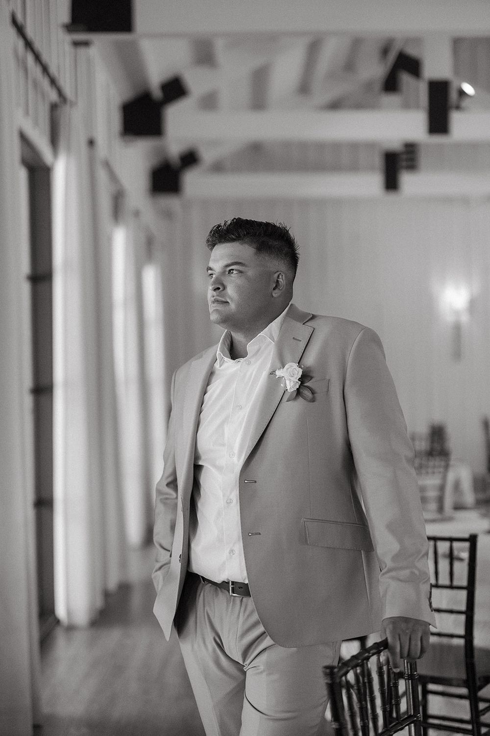 A groom in a light colored suit stands in an elegant white interior venue space.