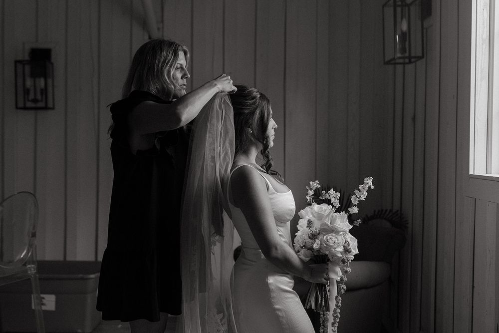 A black and white photograph shows final wedding preparations with a veil being adjusted while holding a floral bouquet.