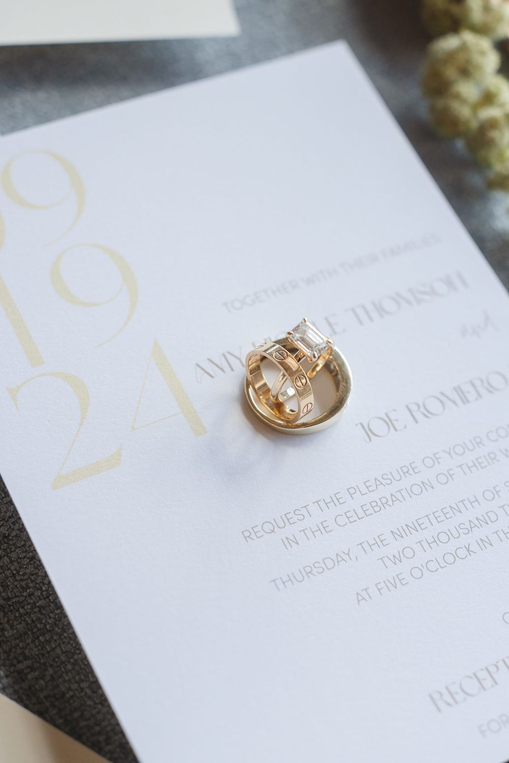 Gold wedding rings rest on top of an elegant white wedding invitation with minimalist typography.