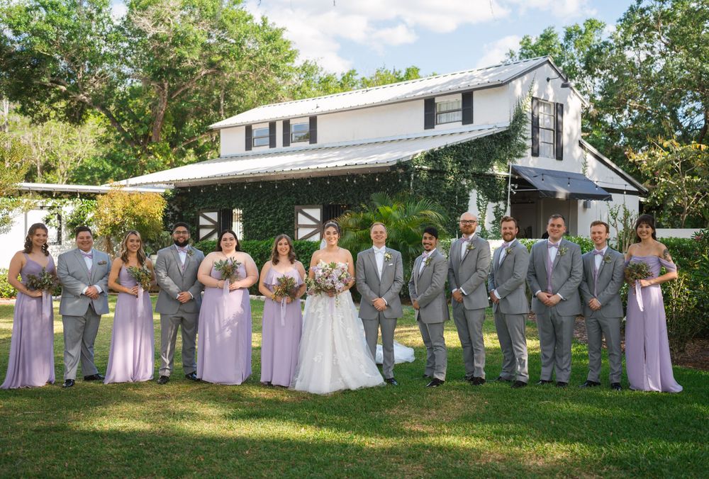 A wedding party in lavender dresses and gray suits poses outdoors on a sunny day, creating a traditional group photo.