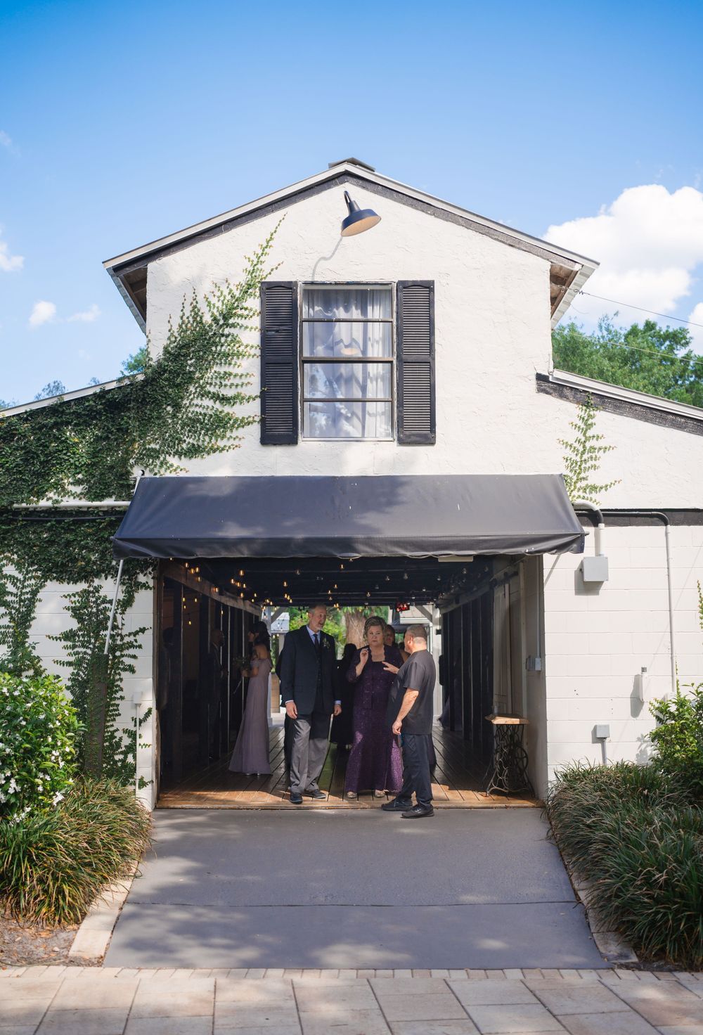 Small white two-story building with black shutters and awning featuring a covered entrance walkway lined with landscaping.