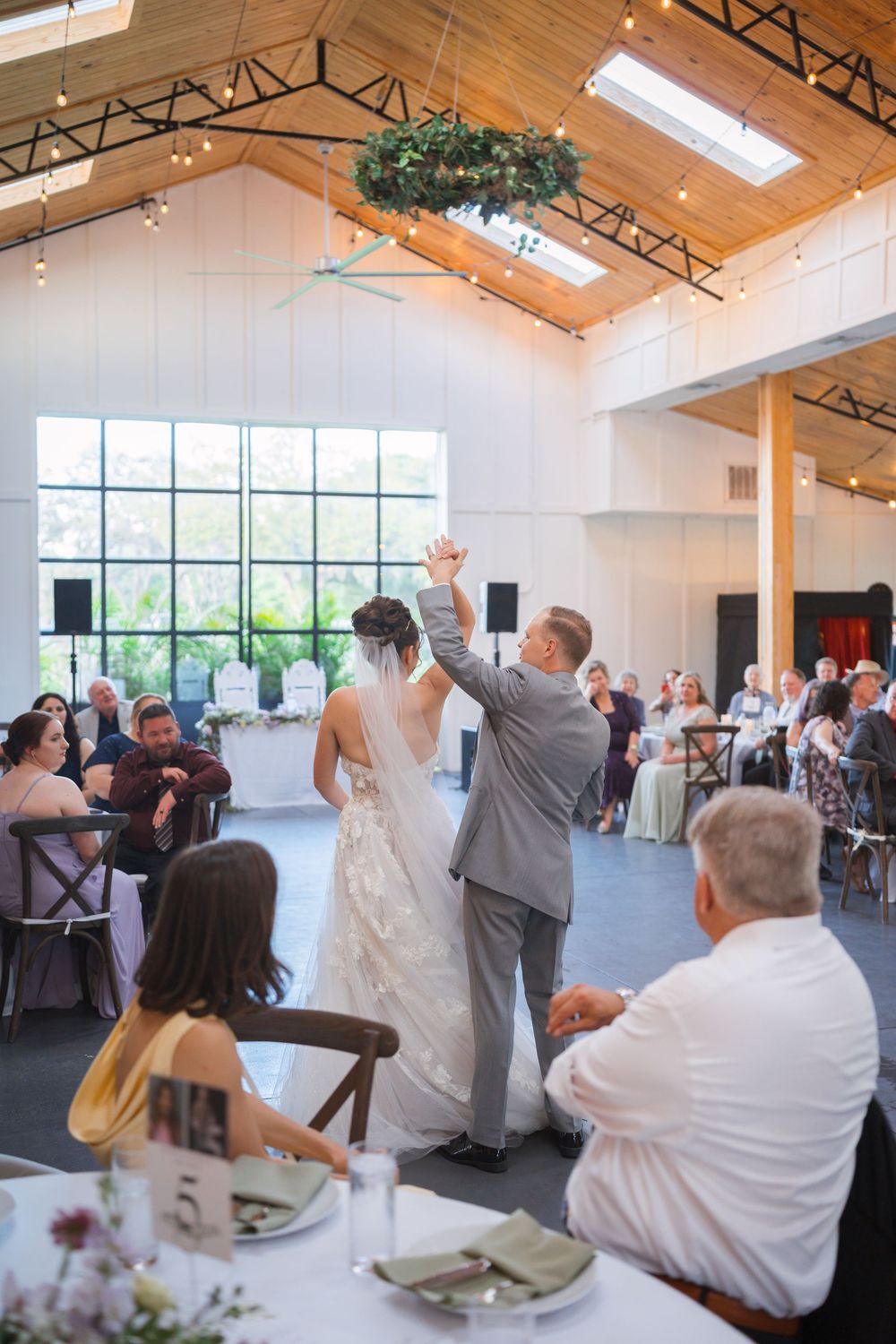 Couple celebrates in joyous moment during wedding reception in modern barn venue with high vaulted ceilings and natural light.