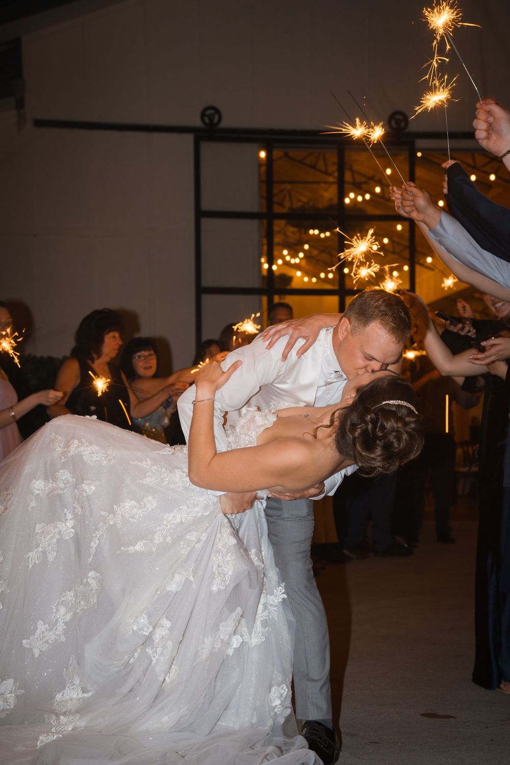A couple shares a romantic dip kiss on their wedding dance floor while guests wave sparklers in celebration.