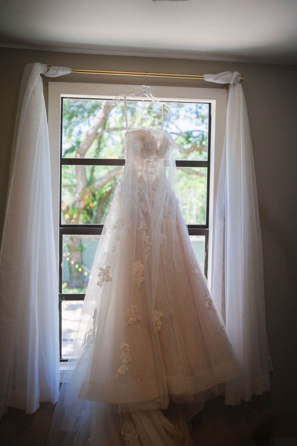 A delicate white wedding dress with floral lace details hangs elegantly in front of a sunlit window with sheer curtains.