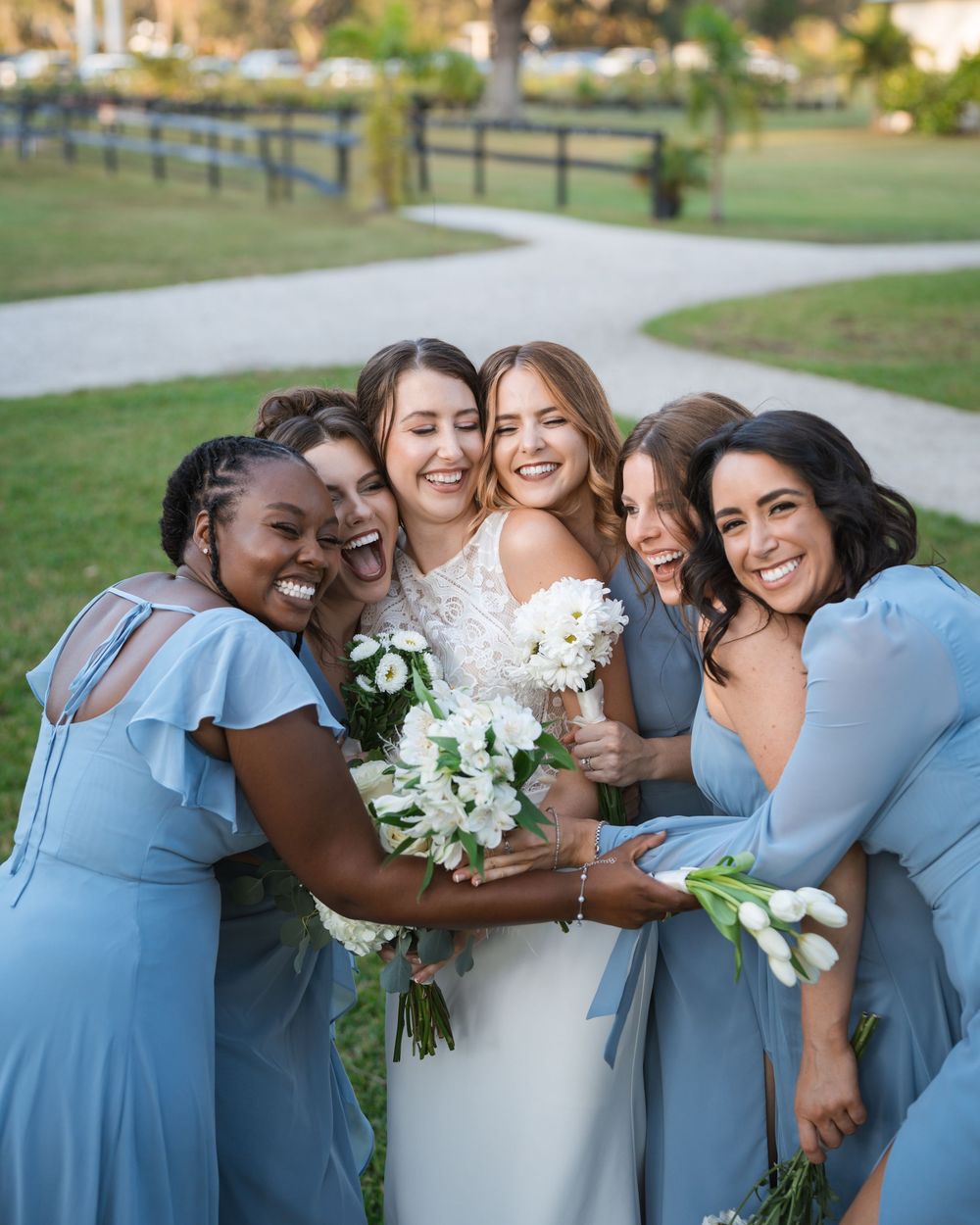 A joyful wedding party wearing light blue bridesmaid dresses share a candid moment of laughter and hugs outside.