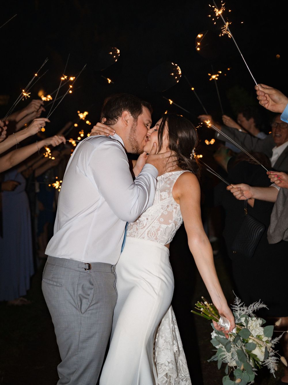A couple shares a romantic kiss under sparklers held by wedding guests during an evening celebration.