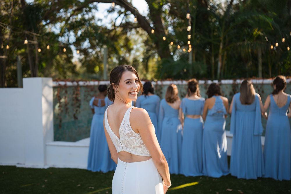 Outdoor wedding scene with bridesmaids in light blue dresses and a smiling bride in a white gown during golden hour.