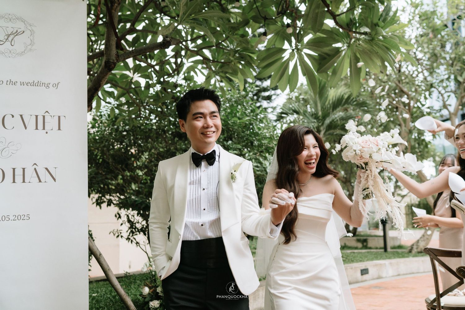 Joyful bride and groom laugh together while walking through outdoor garden wedding venue with lush green trees in background.