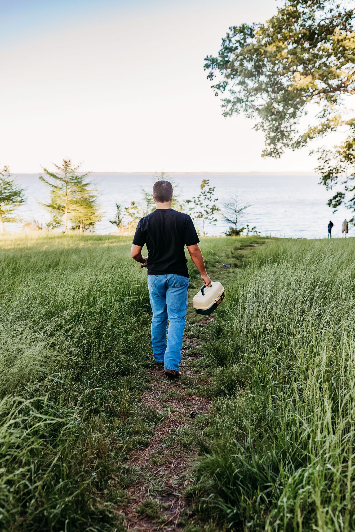 Person walking down a grass trail to the ocean carrying a hat, overlooking coastal waters and evergreen trees at sunset.