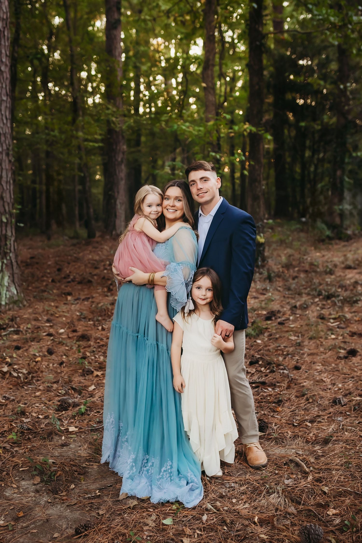 A family poses together in a wooded forest setting wearing coordinated blue and neutral toned outfits.