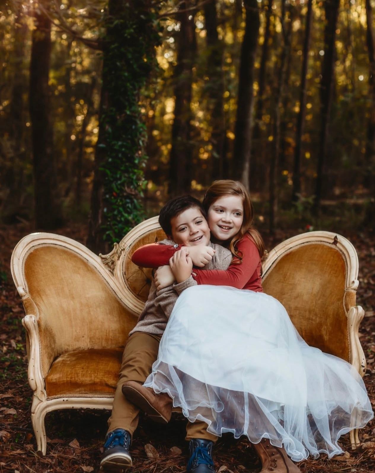 Two children share a sweet moment sitting on a vintage velvet loveseat in an autumn forest with golden sunlight.