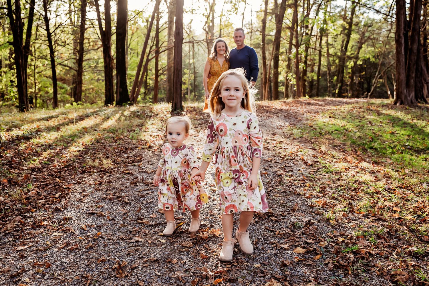 A family walks along a scenic wooded path in autumn with fall leaves scattered on the ground and sunlight filtering through trees.