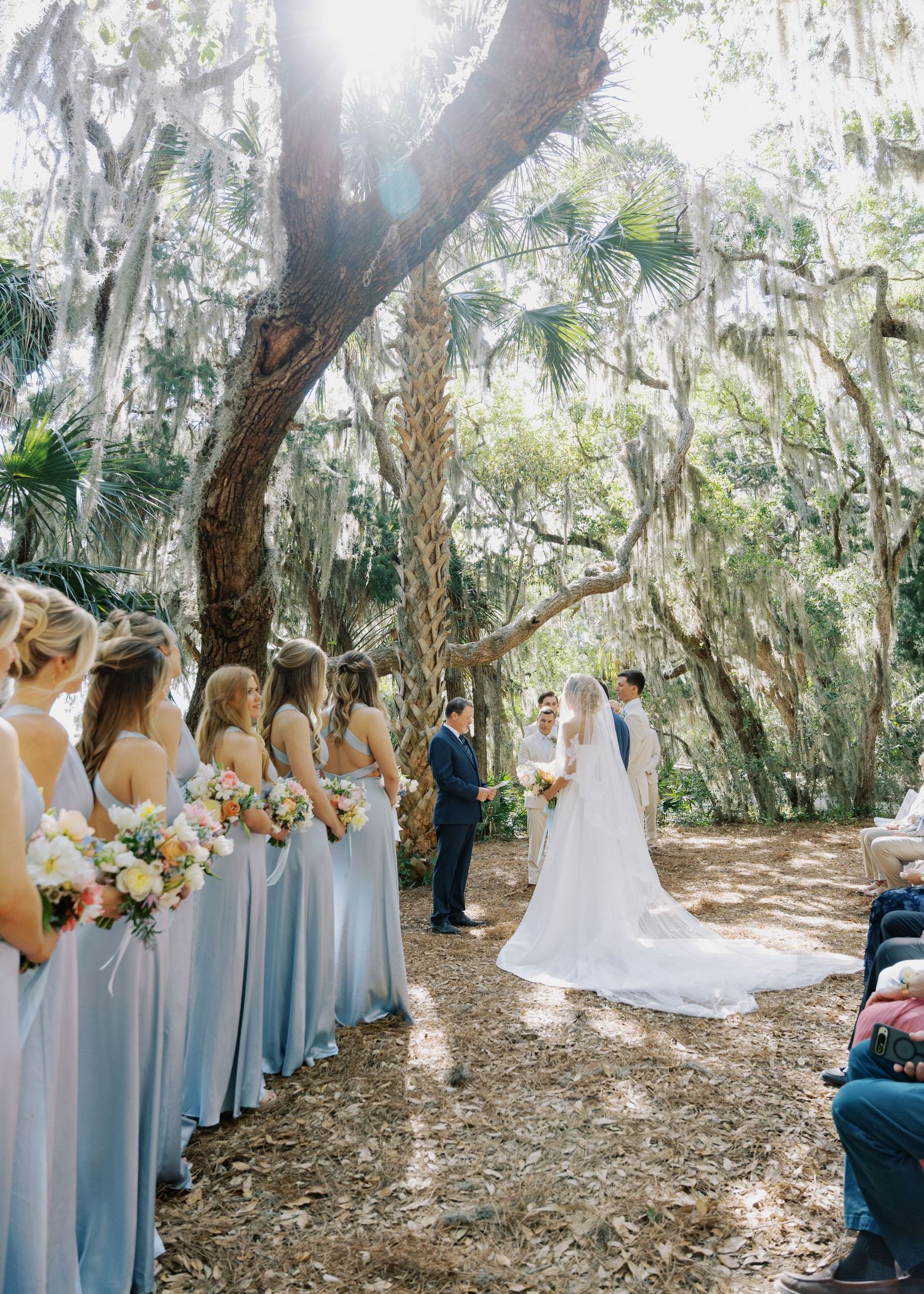 Outdoor wedding ceremony under Spanish moss-draped oak trees with bridesmaids in light blue gowns.
