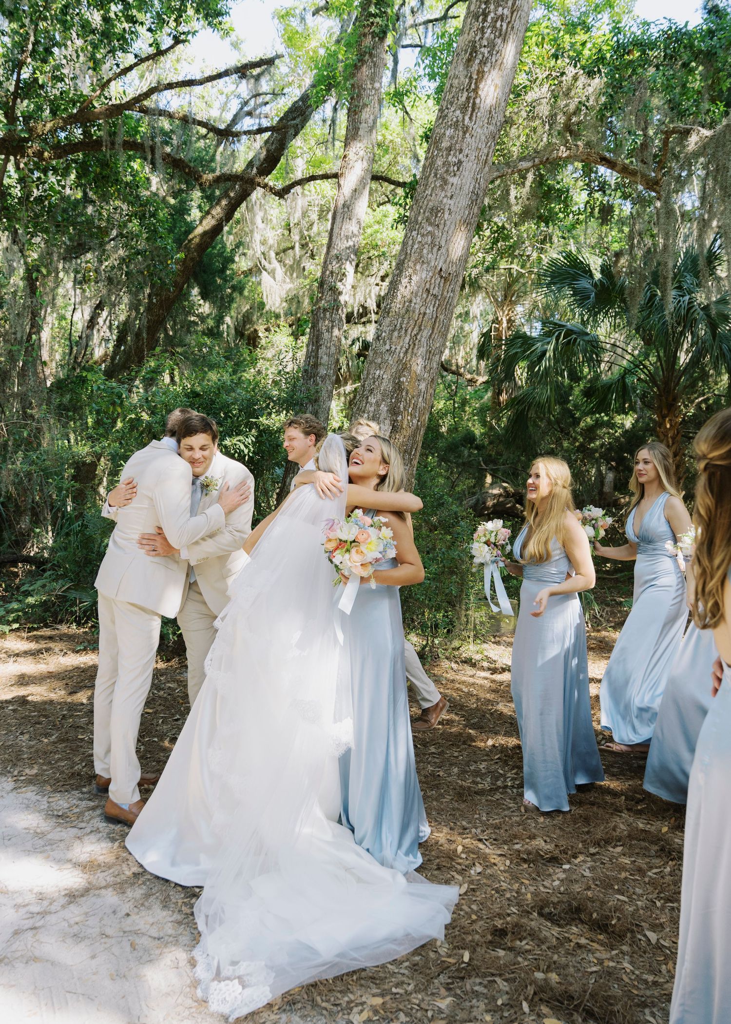Bride and groom share joyful hugs with bridesmaids in light blue gowns among moss-draped oak trees.