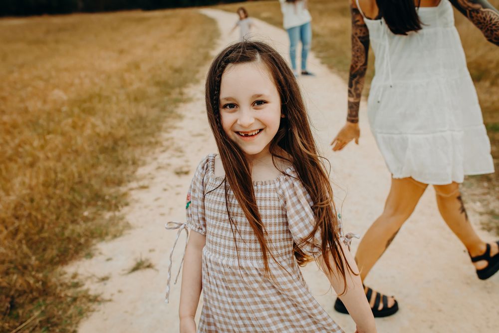 A young person in a gingham dress walks along a path on a sunny day while others follow behind.