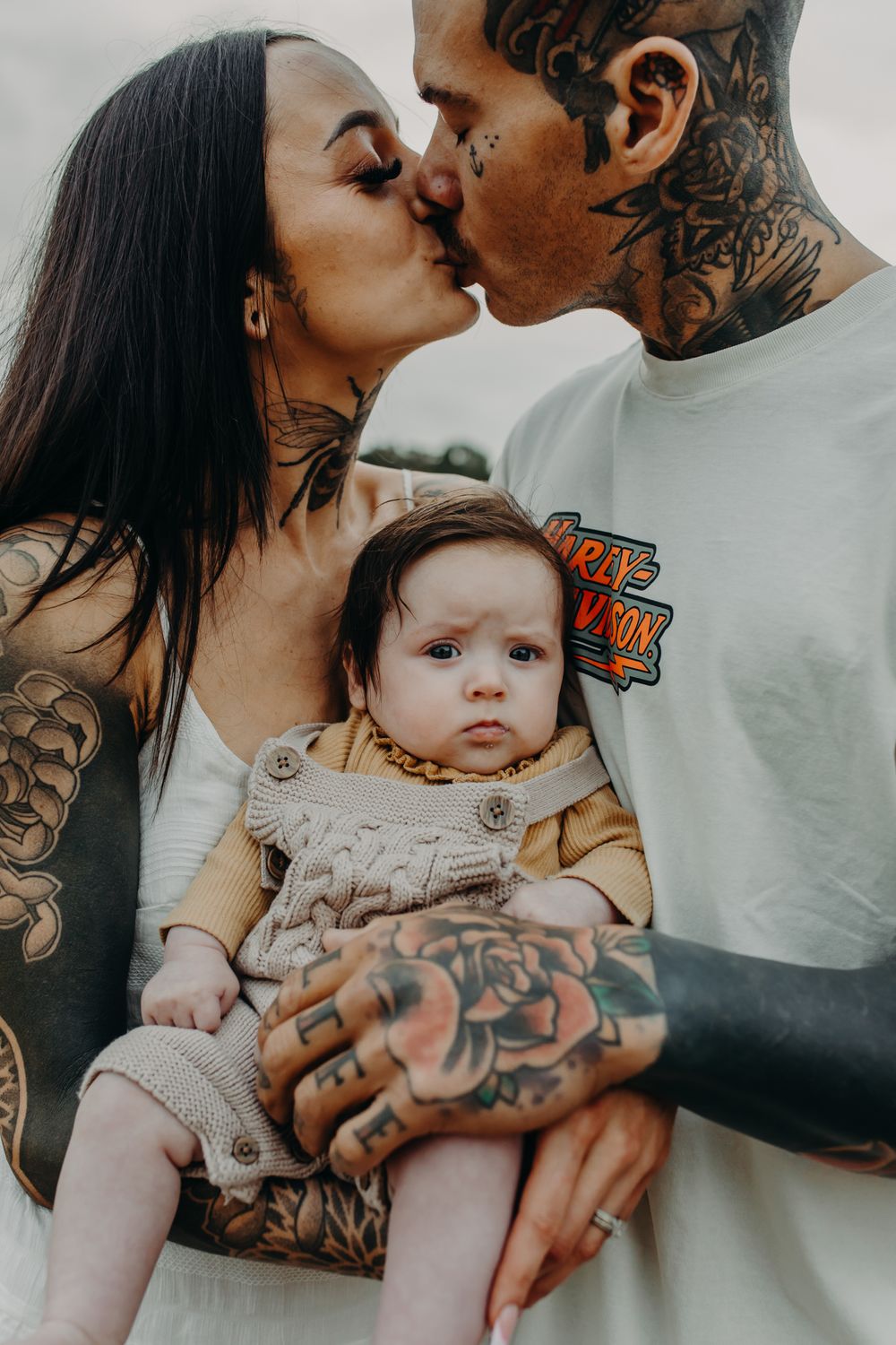 A tattooed couple shares a tender kiss while holding their baby in a heartfelt outdoor portrait.