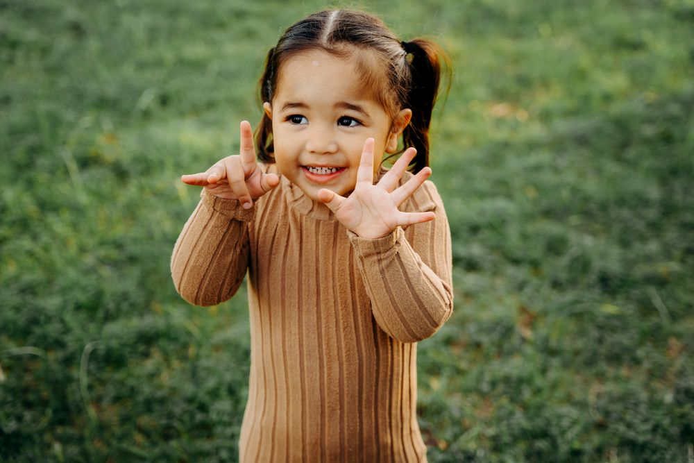 A smiling figure plays with their long brown sweater sleeves against a green grassy background.