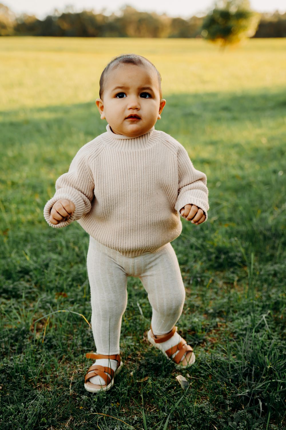 Small child in cream ribbed sweater and matching pants stands on grassy lawn wearing brown leather sandals in golden sunlight.