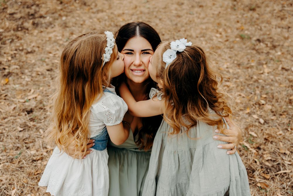 Two children in white dresses giving sweet kisses on the cheeks in a heartwarming outdoor family moment.
