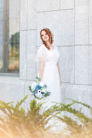Elegant white bridal gown with delicate blue bouquet against stone wall.