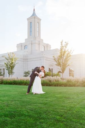 Romantic wedding couple poses on lush green lawn in front of white temple during golden hour sunset.