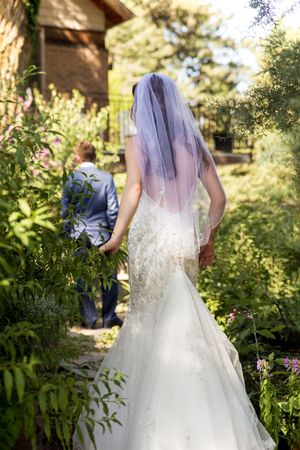 Bride in flowing white gown and veil walks through garden pathway.