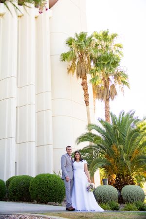 Wedding couple poses against white pillars with tropical palm trees in an elegant outdoor setting.