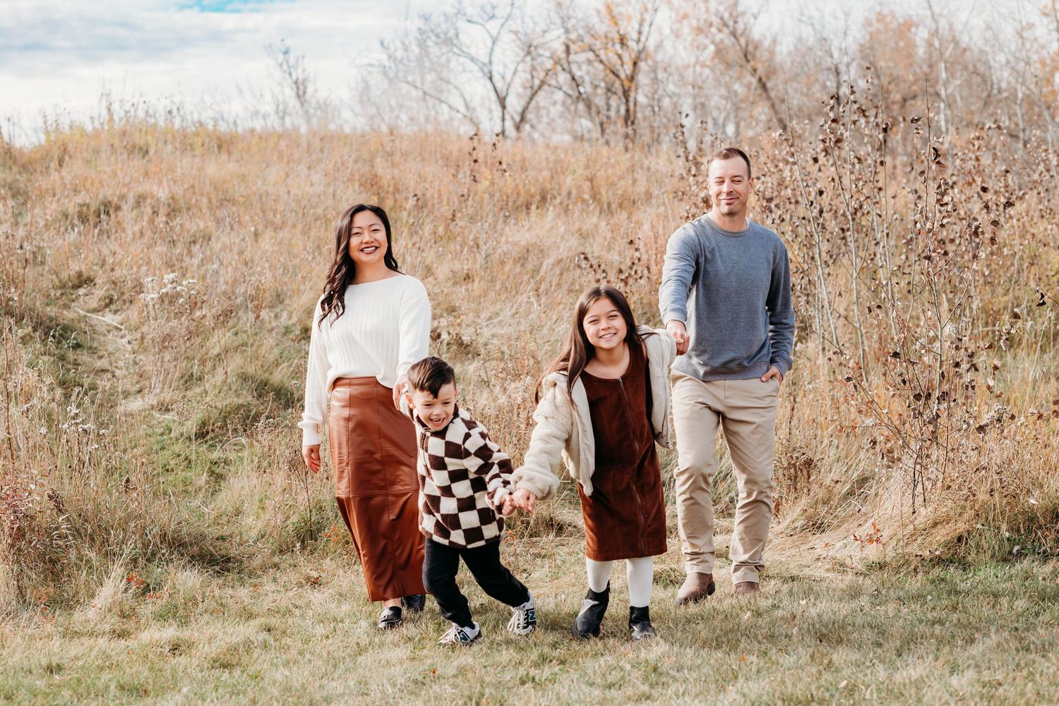 A family of four poses outdoors together on a autumn hillside with tall dried grass in the background.