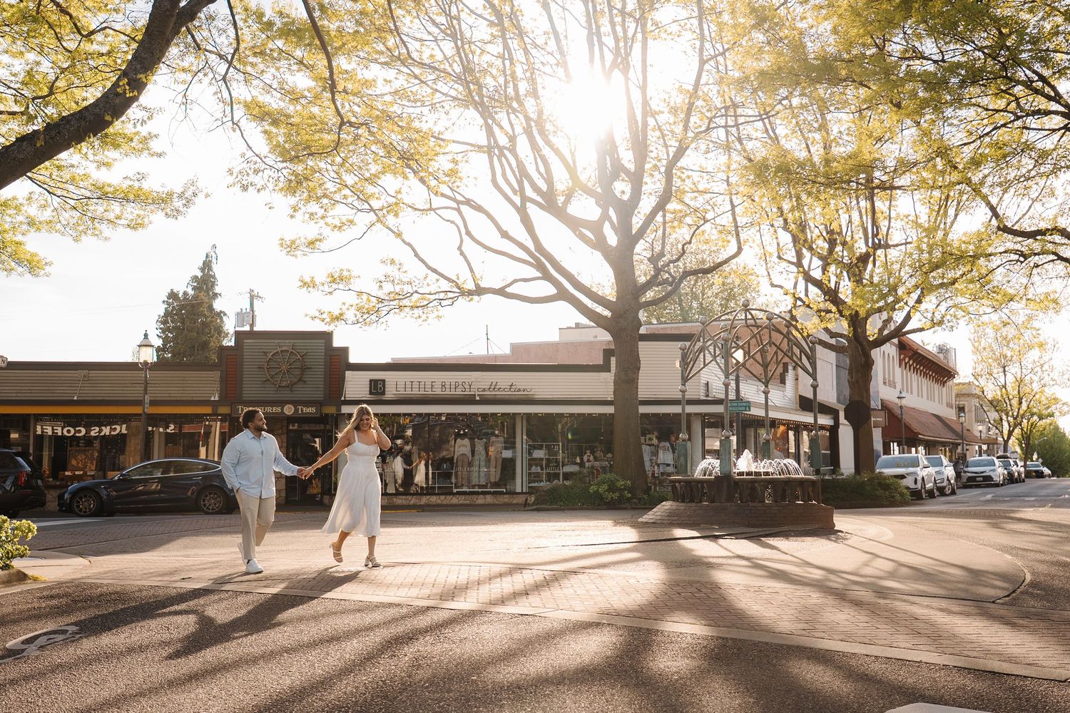 Golden autumn sunlight streams through trees onto a sidewalk with storefronts and people walking in a small town business district.