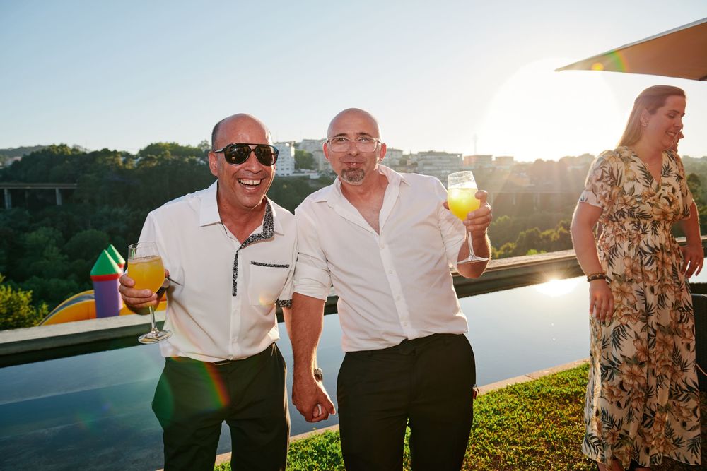 Two smiling men holding colorful cocktails at an outdoor sunset party by an infinity pool.