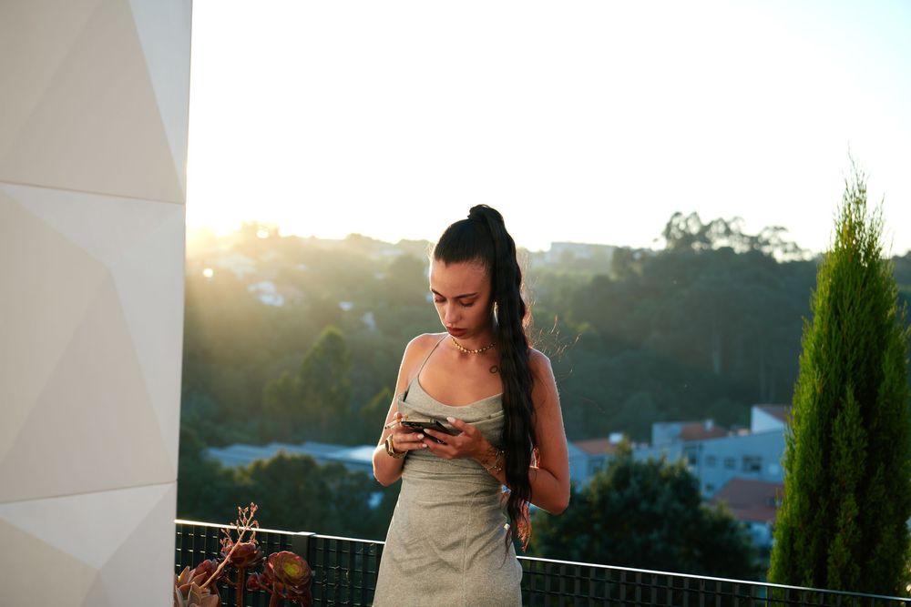 Young woman in a slip dress checking her phone on a sunlit balcony at golden hour.