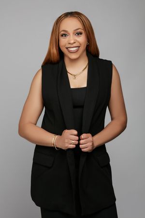 Professional headshot of woman in black sleeveless blazer with gold jewelry against gray background.