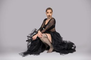 A dancer in a black tulle dress and sheer sleeves poses elegantly against a light gray studio backdrop.