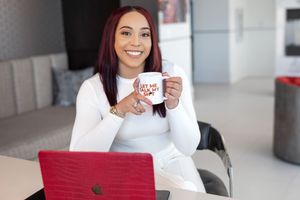 Professional woman in white sweater working on red laptop while holding coffee mug in modern office space.