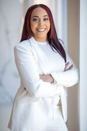 Woman in white blazer stands confidently with arms crossed in bright office setting.