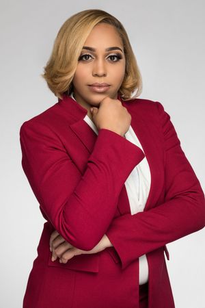 Business professional in red blazer poses with thoughtful expression against grey backdrop.