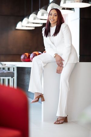 Woman in white business suit sits casually on counter in modern office space.