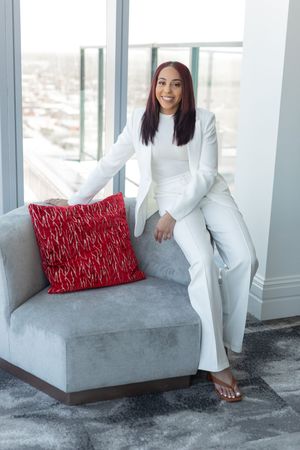 Professional in crisp white suit sits elegantly on gray couch with red accent pillow in modern high-rise space.