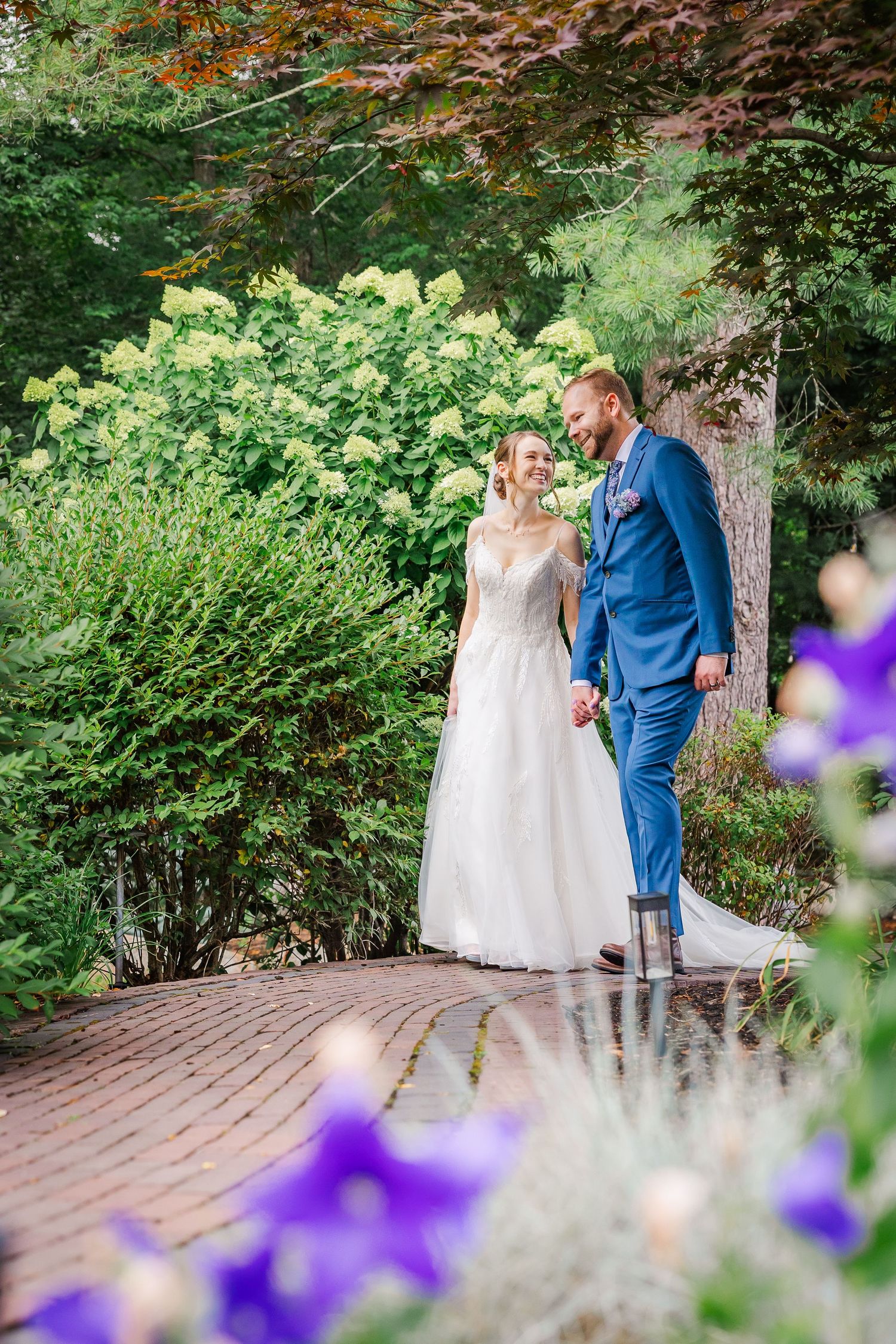 A couple shares an affectionate moment on a brick pathway surrounded by lush garden foliage and purple flowers.